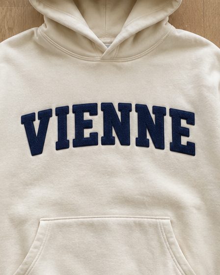 Concept render: cream hoodie with VIENNE in navy chenille letters across the chest, close up of the detail